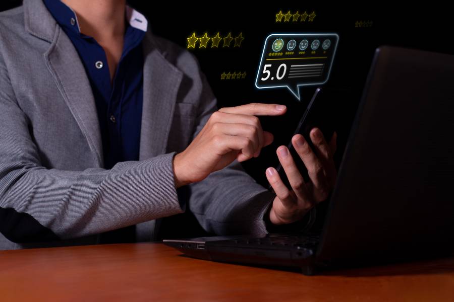 Pessoa segurando smartphone e apontando para uma avaliação digital com nota 5.0 e estrelas, ao lado de um notebook, em pesquisa de satisfação do cliente