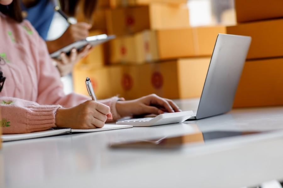 Vendedora anotando em caderno com laptop e caixas ao fundo, gerenciando pontos de penalidade da Shopee